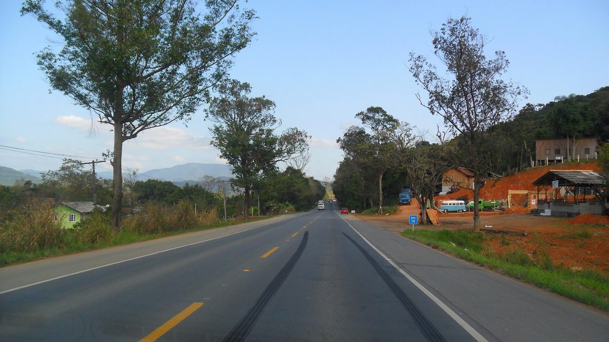 Rodovia de pista simples entre árvores e morros, com placa do km 76, em dia de sol