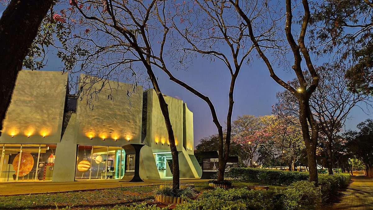 Prédio do Planetário de Brasília iluminado ao entardecer, entre árvores do Eixo Monumental