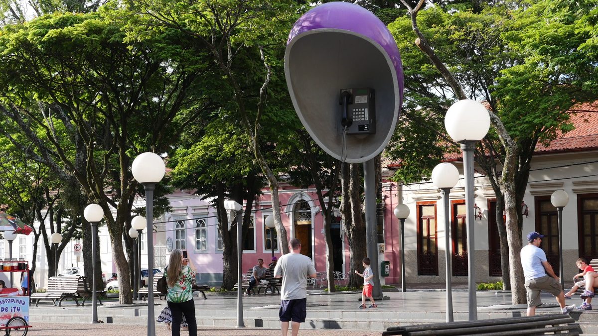 Orelhão gigante na Praça Padre Miguel, no centro de Itu, entre árvores e postes