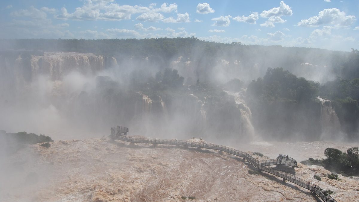 Cataratas do Iguaçu com água barrenta em grande volume e passarela sobre o rio