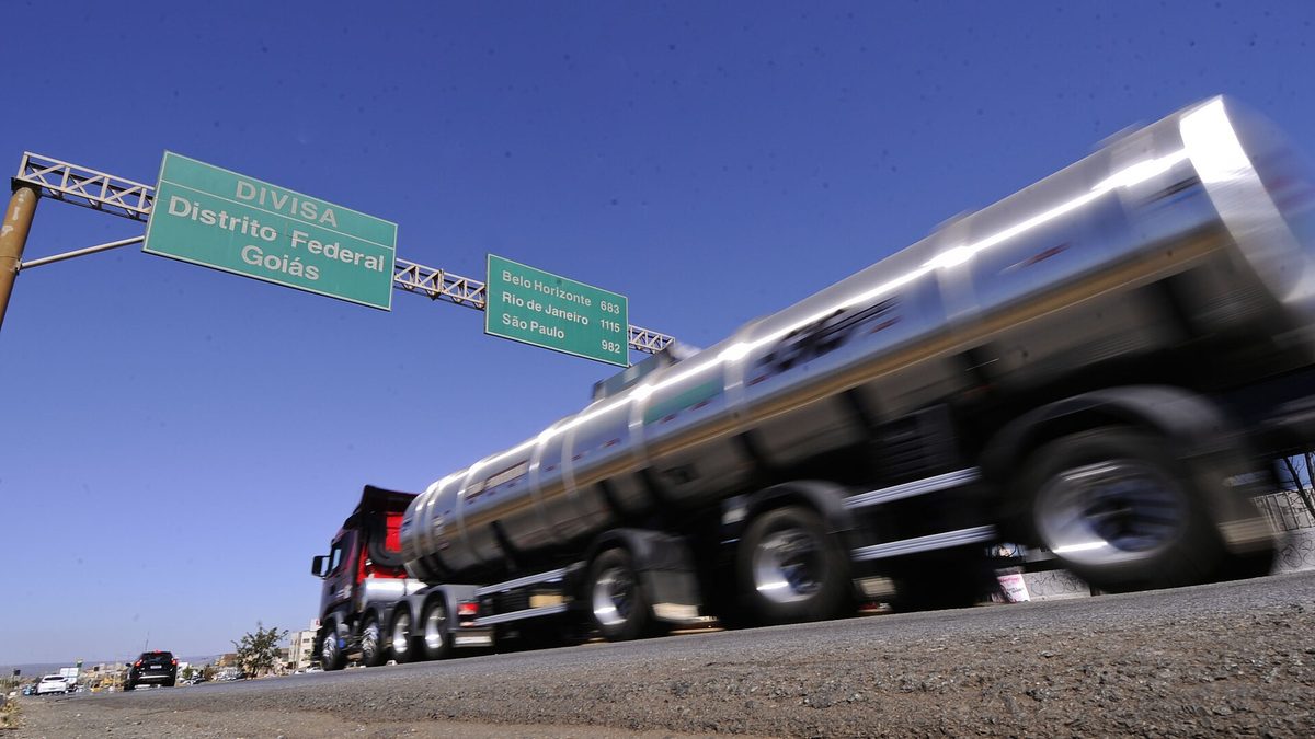 Caminhão-tanque passa sob placas verdes da divisa entre o Distrito Federal e Goiás, com céu azul