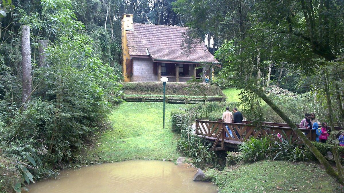 Casa de madeira e ponte sobre lago no Bosque Alemão, em Curitiba, com visitantes