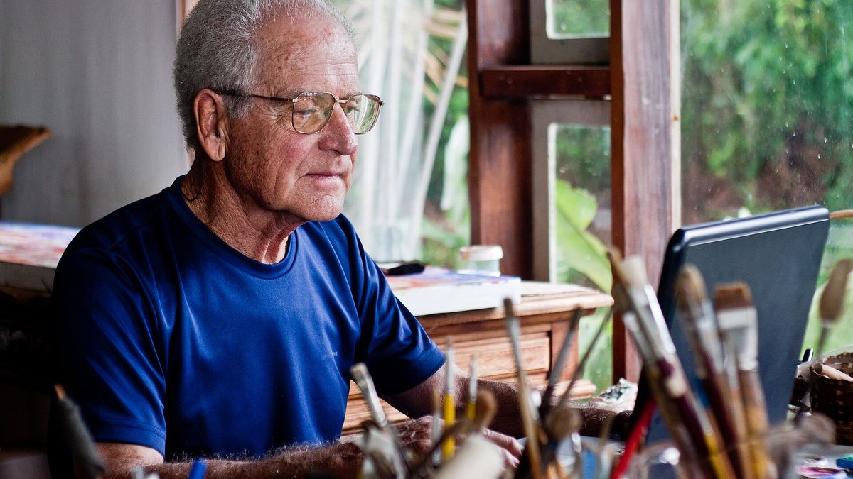 Homem idoso de óculos usa um notebook em ateliê, com pincéis em primeiro plano