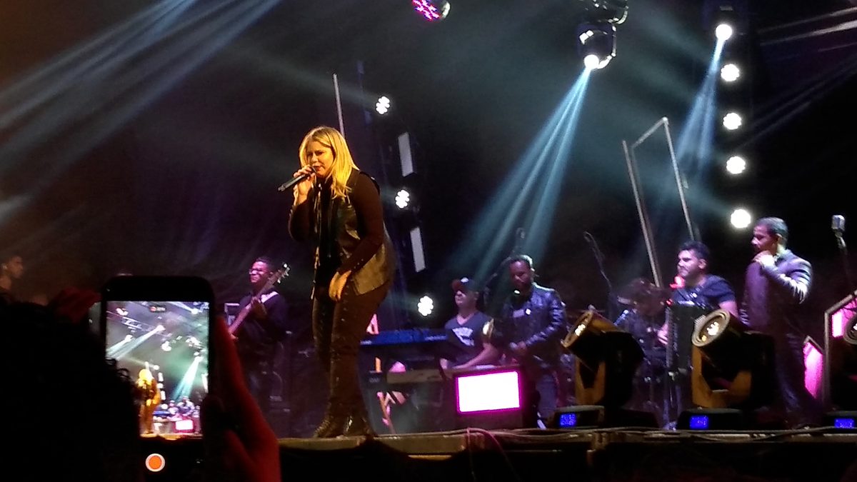 Marília Mendonça canta em show, com a banda ao fundo e luzes azuis no palco