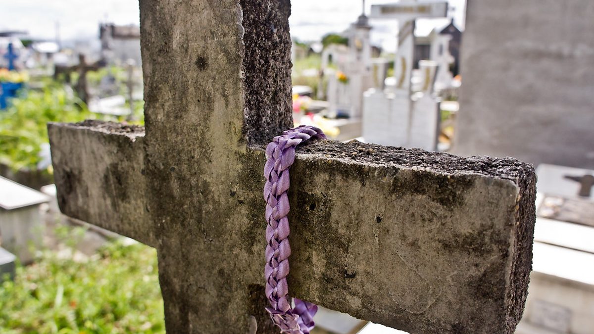 Cruz de pedra com uma fita roxa amarrada em cemitério, com túmulos brancos ao fundo