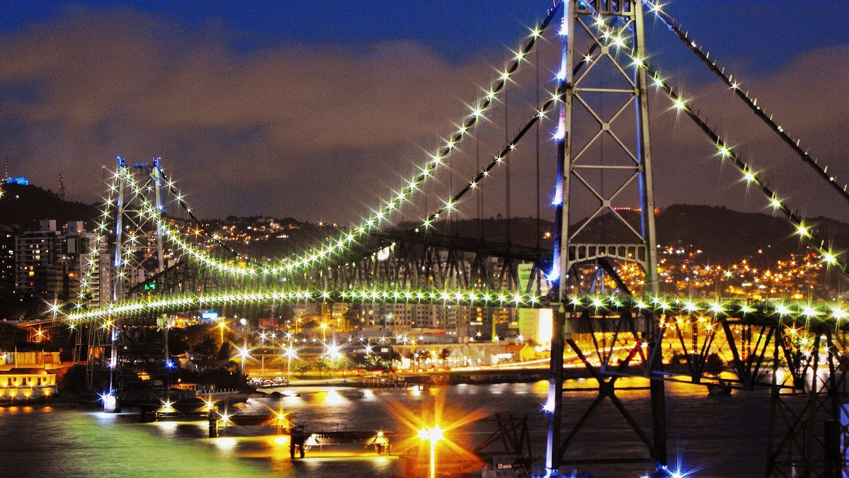 Ponte Hercílio Luz iluminada à noite, com o Centro de Florianópolis ao fundo