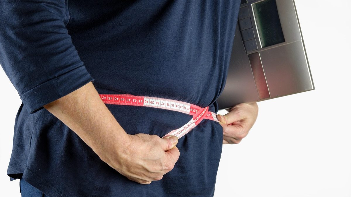 Pessoa mede a cintura com fita métrica por cima da camiseta, segurando uma balança