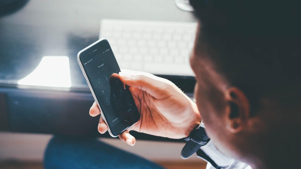 Homem digita a senha na tela de bloqueio do celular, diante do teclado do computador