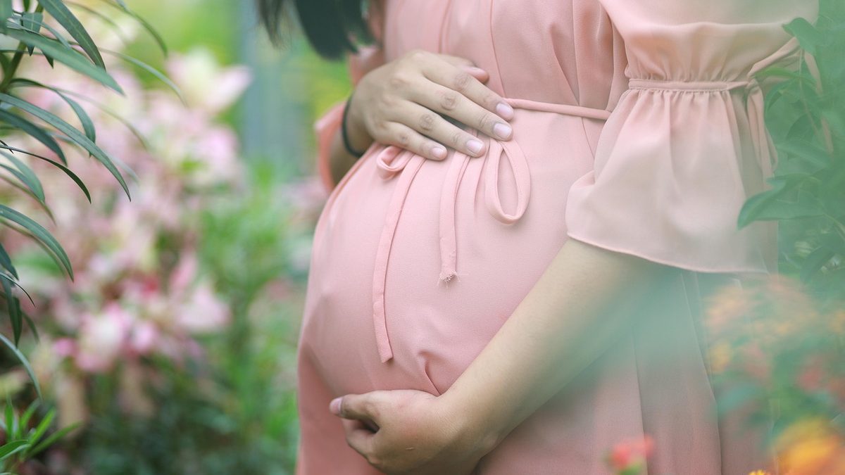 Gestante de vestido rosa com as mãos sobre a barriga, entre flores