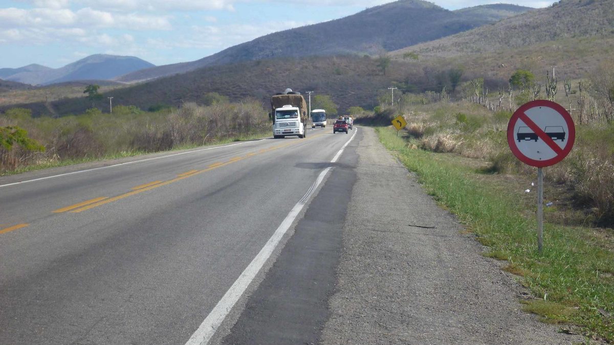 Pista simples da BR-116 entre morros na Bahia, com caminhão, ônibus e placa de proibido ultrapassar