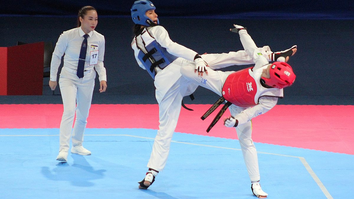 Maria Clara Pacheco e Chaima Toumi trocam chutes em combate de taekwondo, sob o olhar da árbitra
