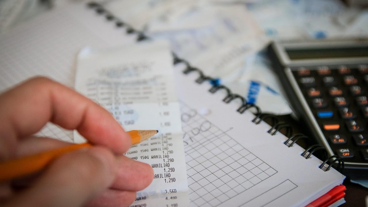 Mão com lápis confere um cupom de compras ao lado de um caderno e de uma calculadora