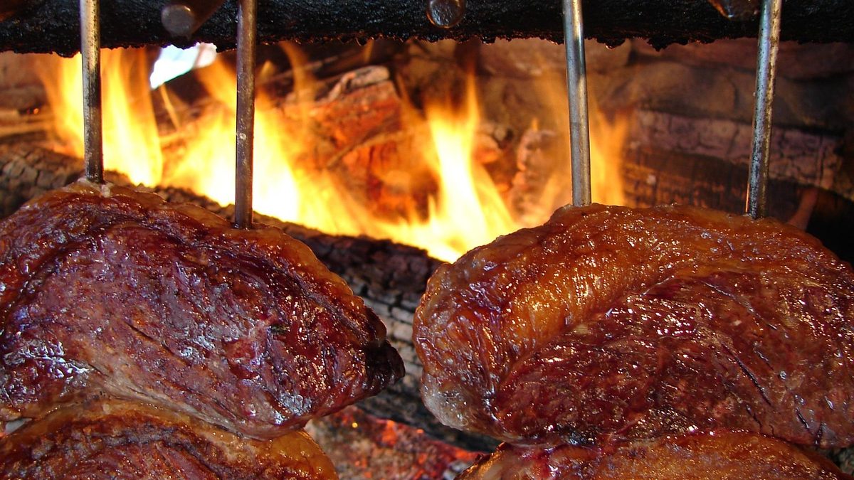 Peças de carne assando em espetos diante do fogo de uma churrasqueira