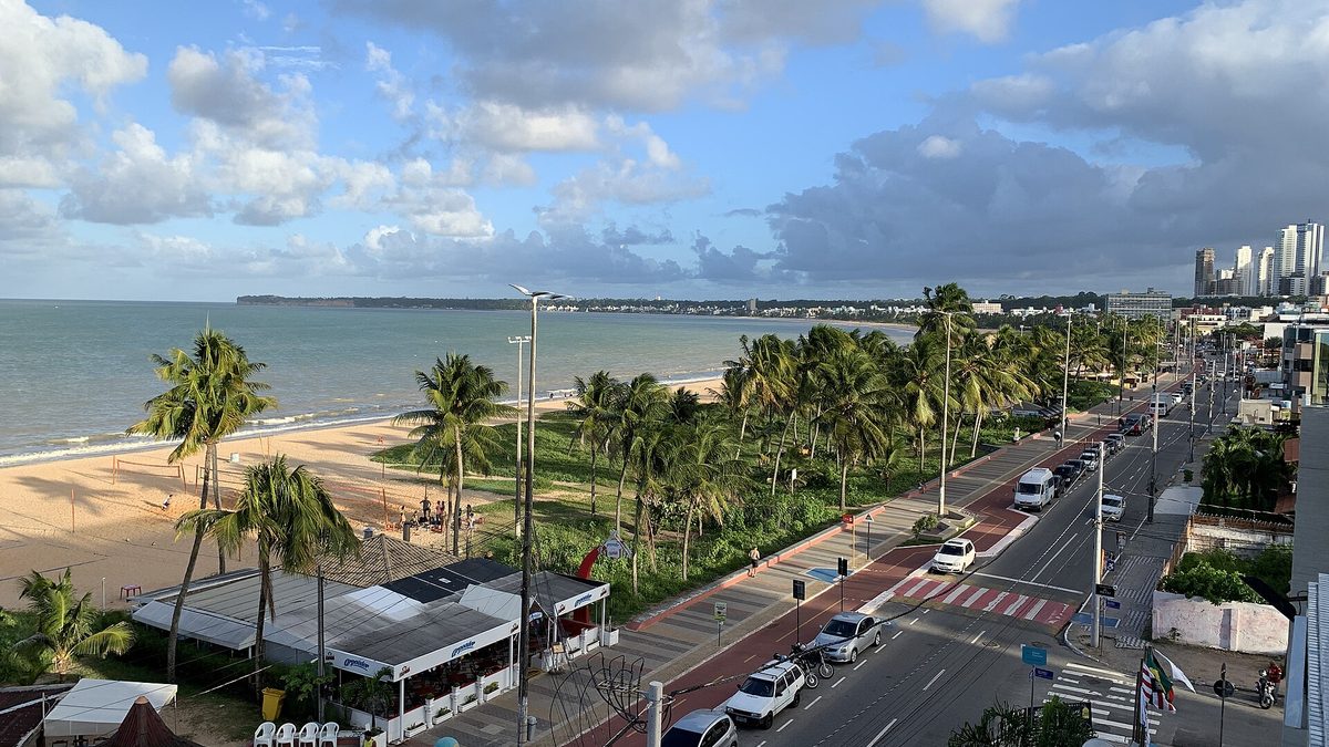 Avenida à beira-mar com coqueiros, calçadão e prédios ao fundo, na orla de João Pessoa