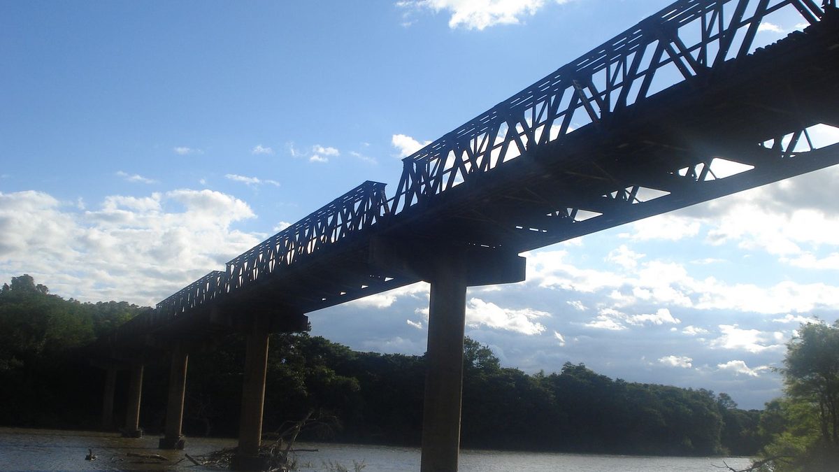 Ponte metálica de treliça sobre o Rio Vacacaí, com mata nas margens e céu azul