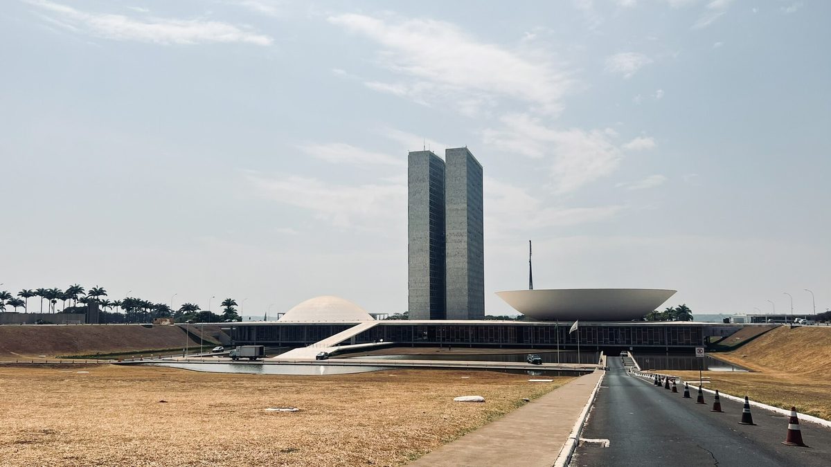 Congresso Nacional com gramado seco sob céu esbranquiçado, em Brasília