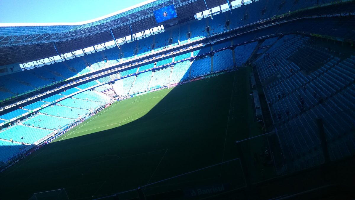 Arquibancadas azuis e gramado da Arena do Grêmio vistos de dentro do estádio, em Porto Alegre