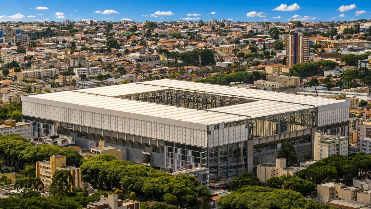 Vista aérea da Arena da Baixada num dia de sol, com cobertura branca, árvores verdes e céu azul, em Curitiba