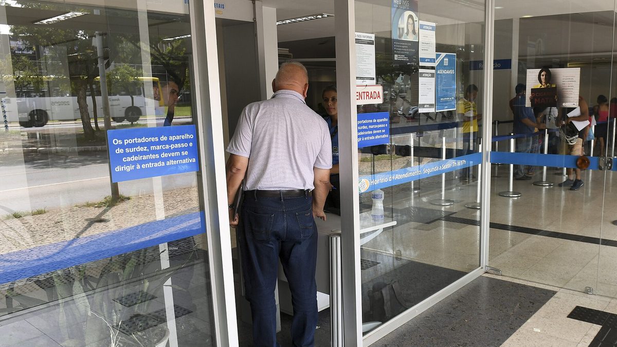 Segurado de costas entra pela porta de vidro de uma agência do INSS