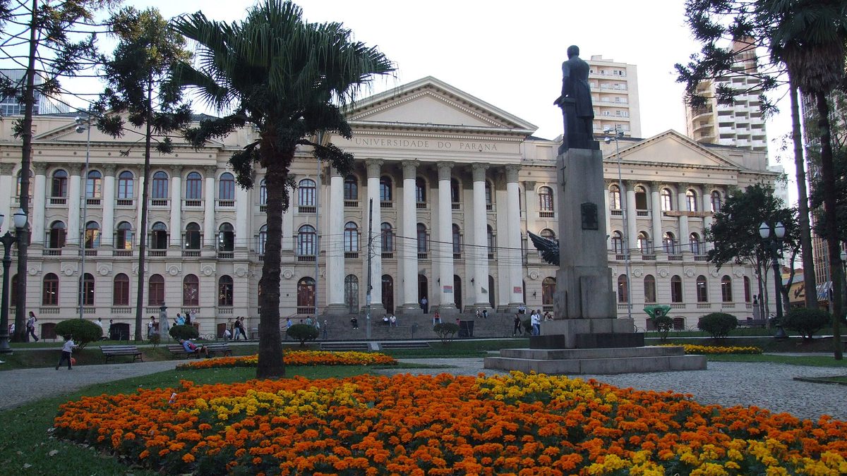 Prédio histórico da UFPR com colunas brancas e canteiro de flores na Praça Santos Andrade, em Curitiba
